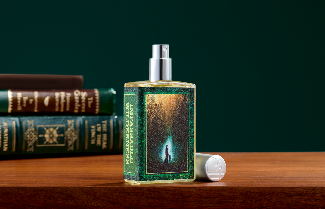 A rectangular glass perfume bottle labeled 'Essential Wilderness' with a silver spray top, standing on a wooden surface. A matching white cap lies beside it, and stacked books are blurred in the background against a dark green backdrop.