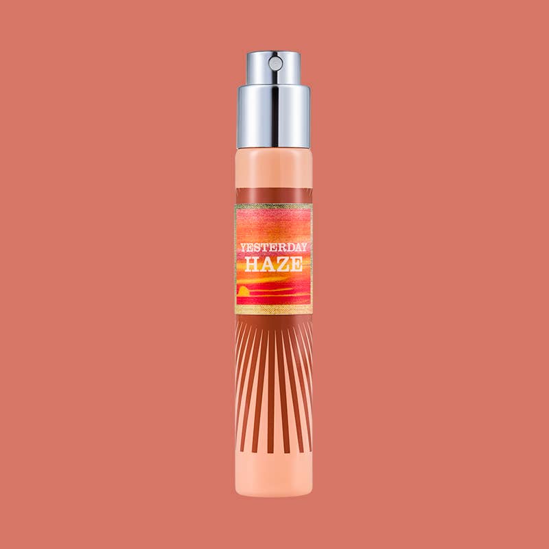 A single cylindrical spray bottle of 'Yesterday Haze' fragrance, featuring a peach-colored body with a silver pump and a colorful label, centered on a solid terracotta background.